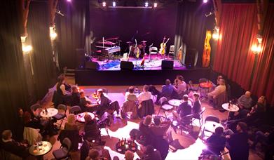 People sitting at individual tables in front of the stage at the Chapel Arts Centre in Bath